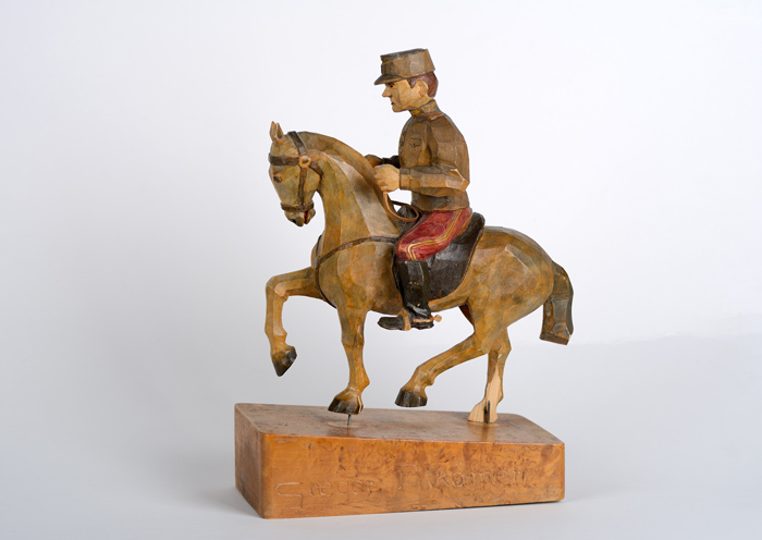 Kuvassa on puusta veistetty pieni patsas, joka esittää punahousuista rakuunasotilasta hevosen selässä.